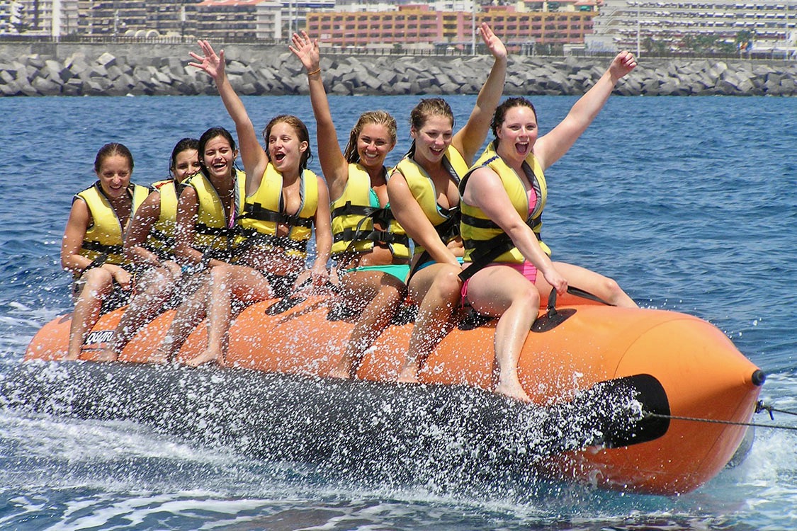 Un grupo de niñas saludando y riendo mientras su banana boat avanza a toda velocidad por el agua.